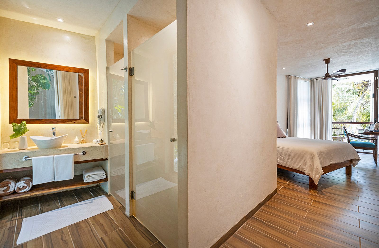 Bathroom and bedroom with shower, sink, mirror, and bed. Light-colored walls and wood flooring.