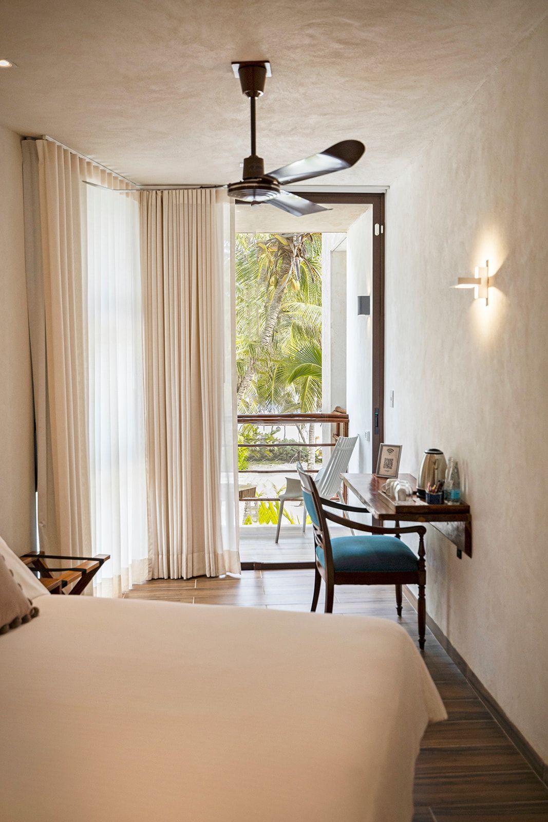 Bedroom with bed, desk, and balcony. Soft light, neutral tones, and a ceiling fan.
