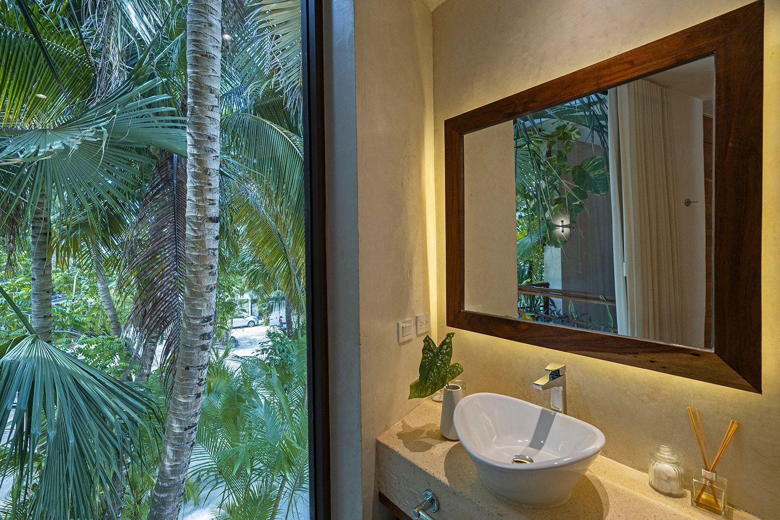 Bathroom with large window overlooking lush trees; white sink with mirror and accent lighting.