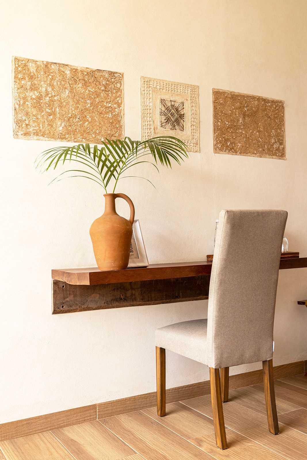Desk with a clay vase holding greenery, three textured art pieces, and a gray chair against a white wall.