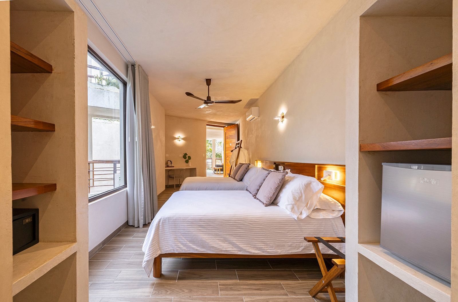 Bedroom with two beds, built-in shelves, large window, neutral colors, and a view of an outdoor area.