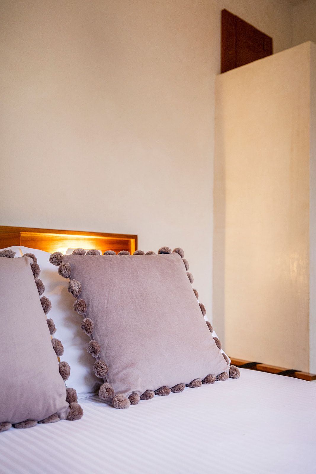 Two square pillows with pom-pom trim on a white bed. Beige walls and a wooden headboard are behind.