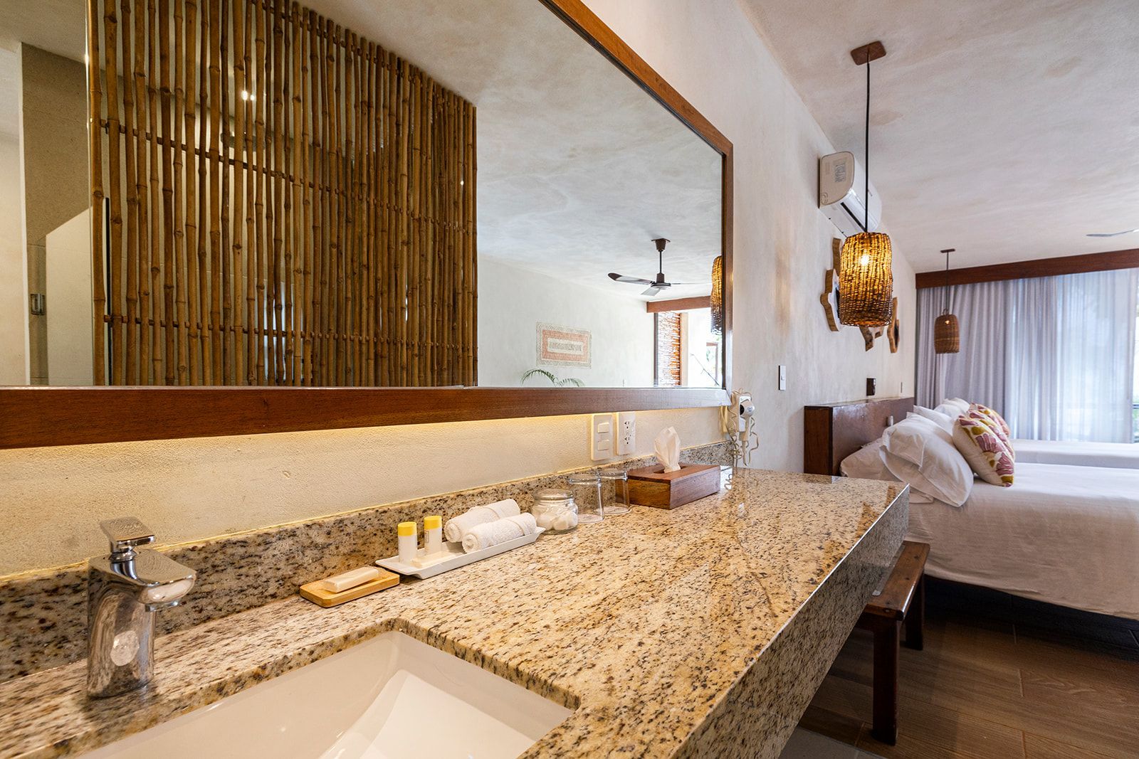 Bathroom with granite countertop, large mirror, bamboo wall, and a bed in the background.