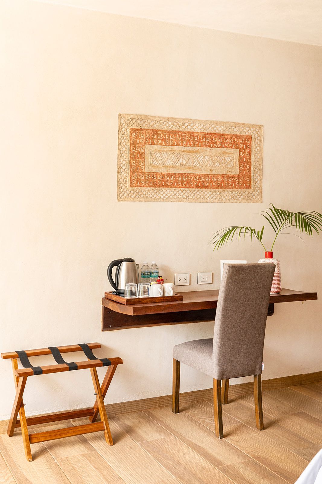 Desk area with floating shelf, chair, luggage rack. Neutral wall with decorative wall hanging and wood flooring.