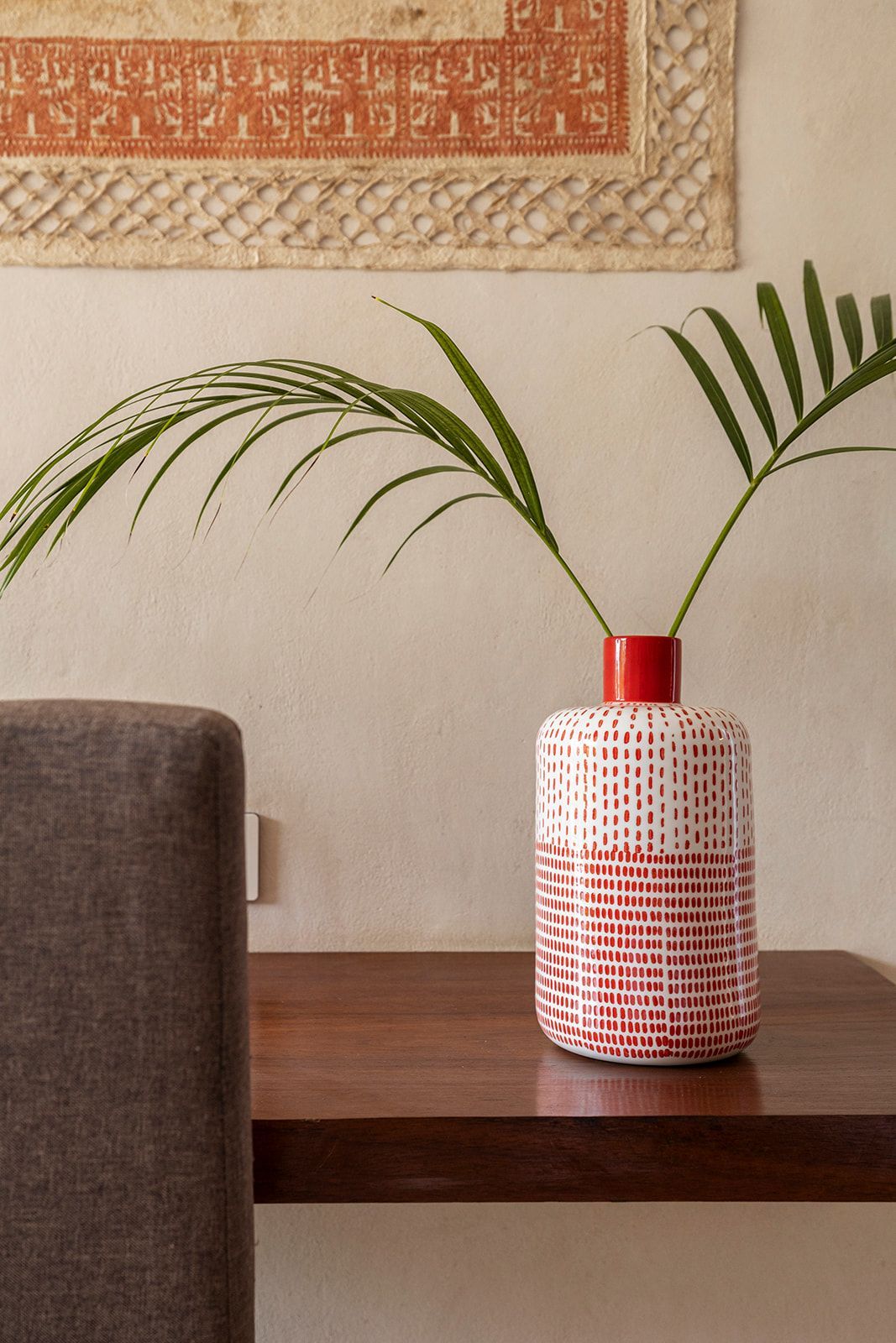 Red and white patterned vase with green palm fronds on a dark wood table. Wall with textured tapestry.