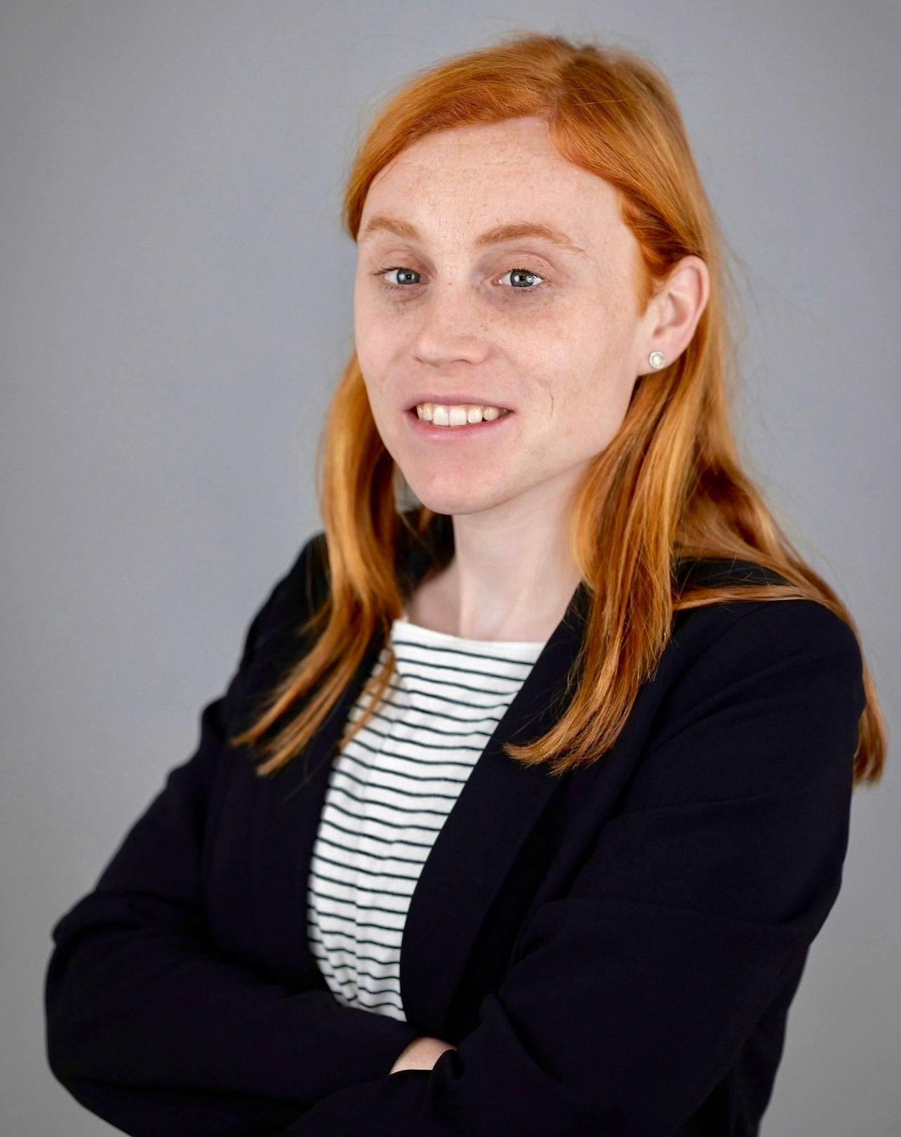 Woman with red hair, wearing a striped shirt and blazer, arms crossed, smiling.