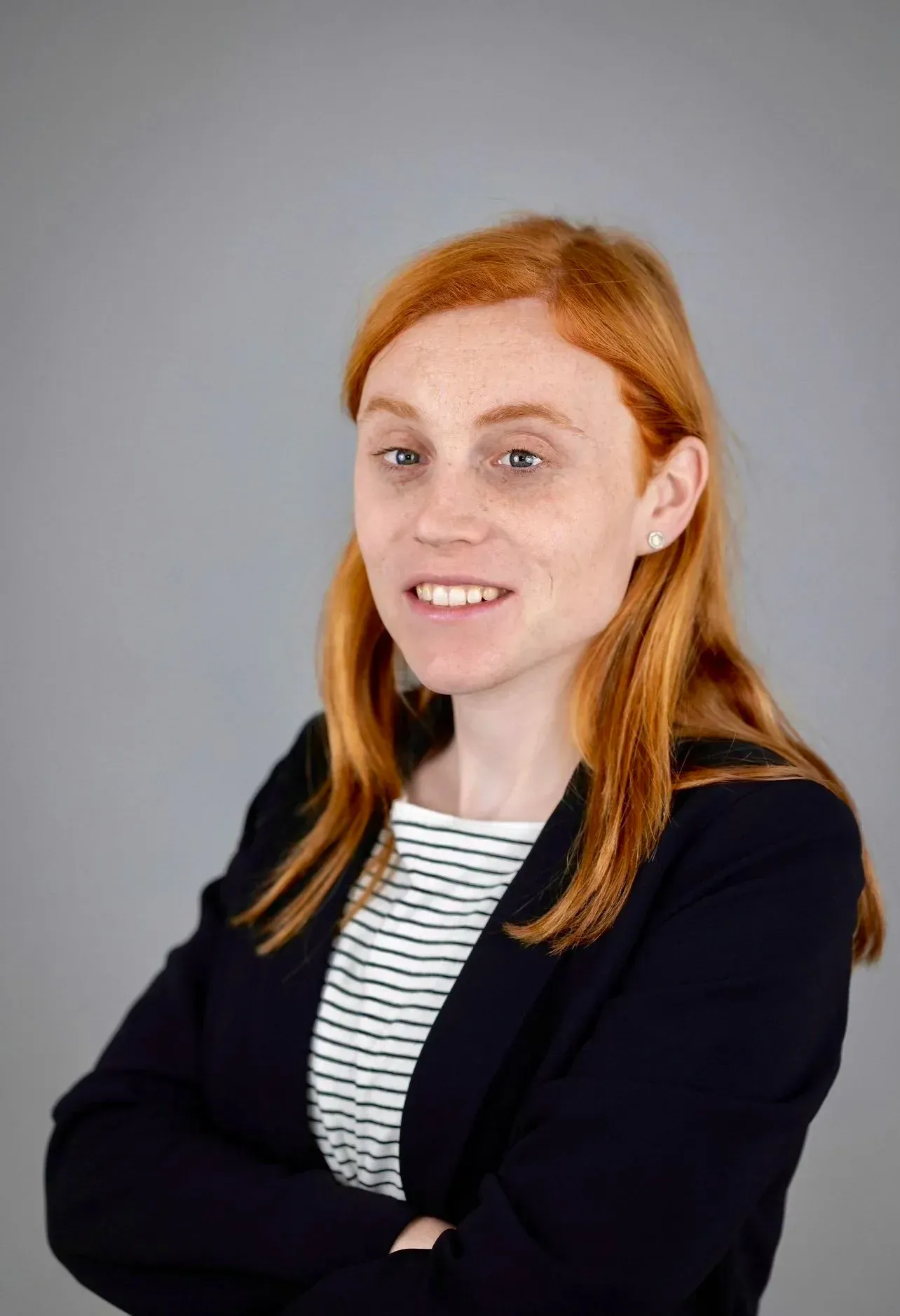 A person with red hair and freckles, wearing a black blazer over a striped top, smiling against a plain gray background.