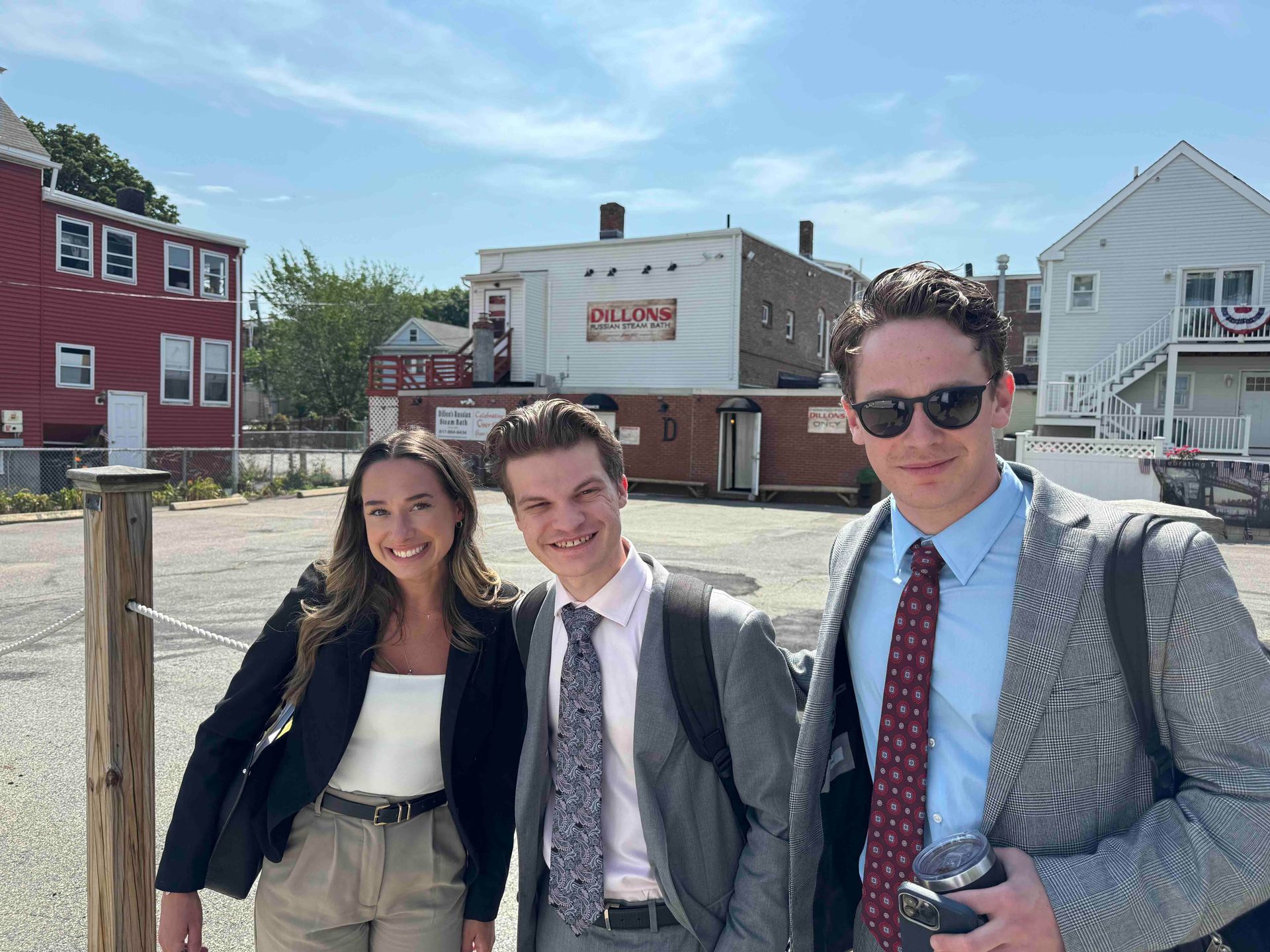 Three people stand smiling in an outdoor parking lot in front of a white building on a sunny day.