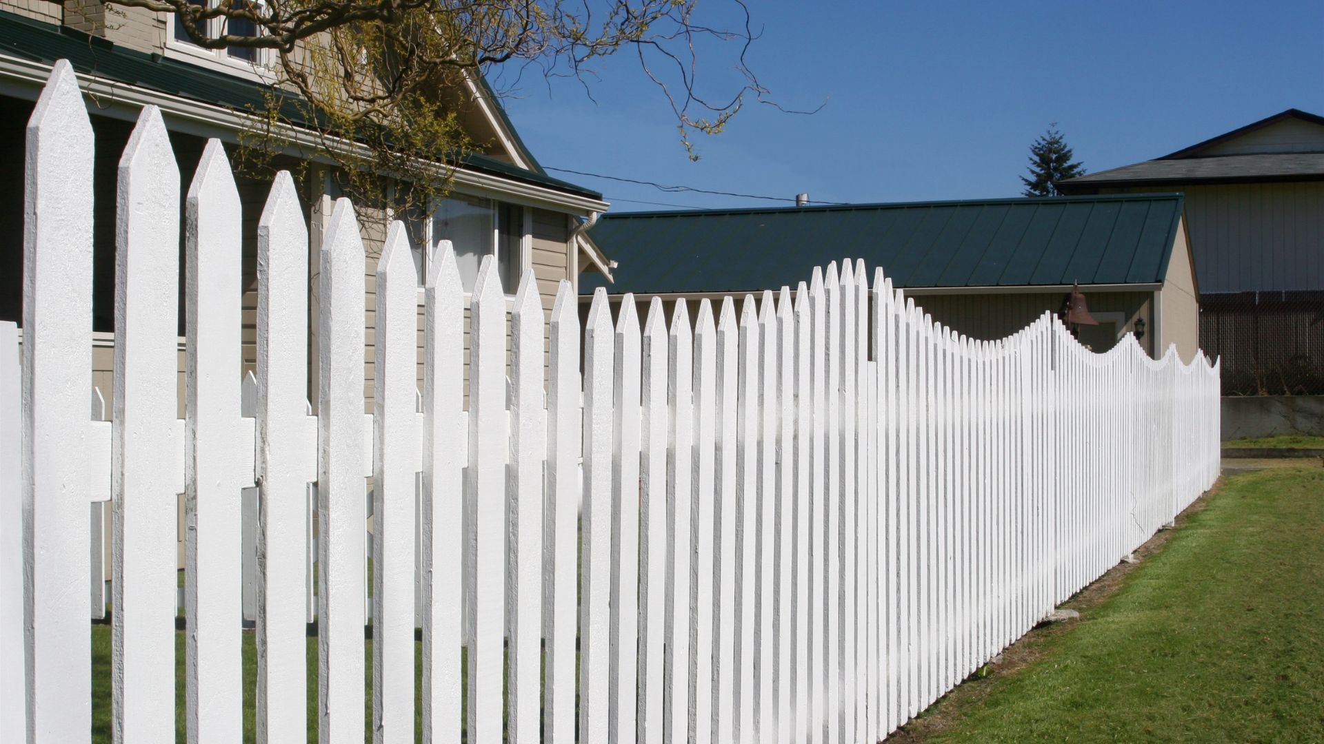 White picket fence beside a house and grassy yard under a blue sky