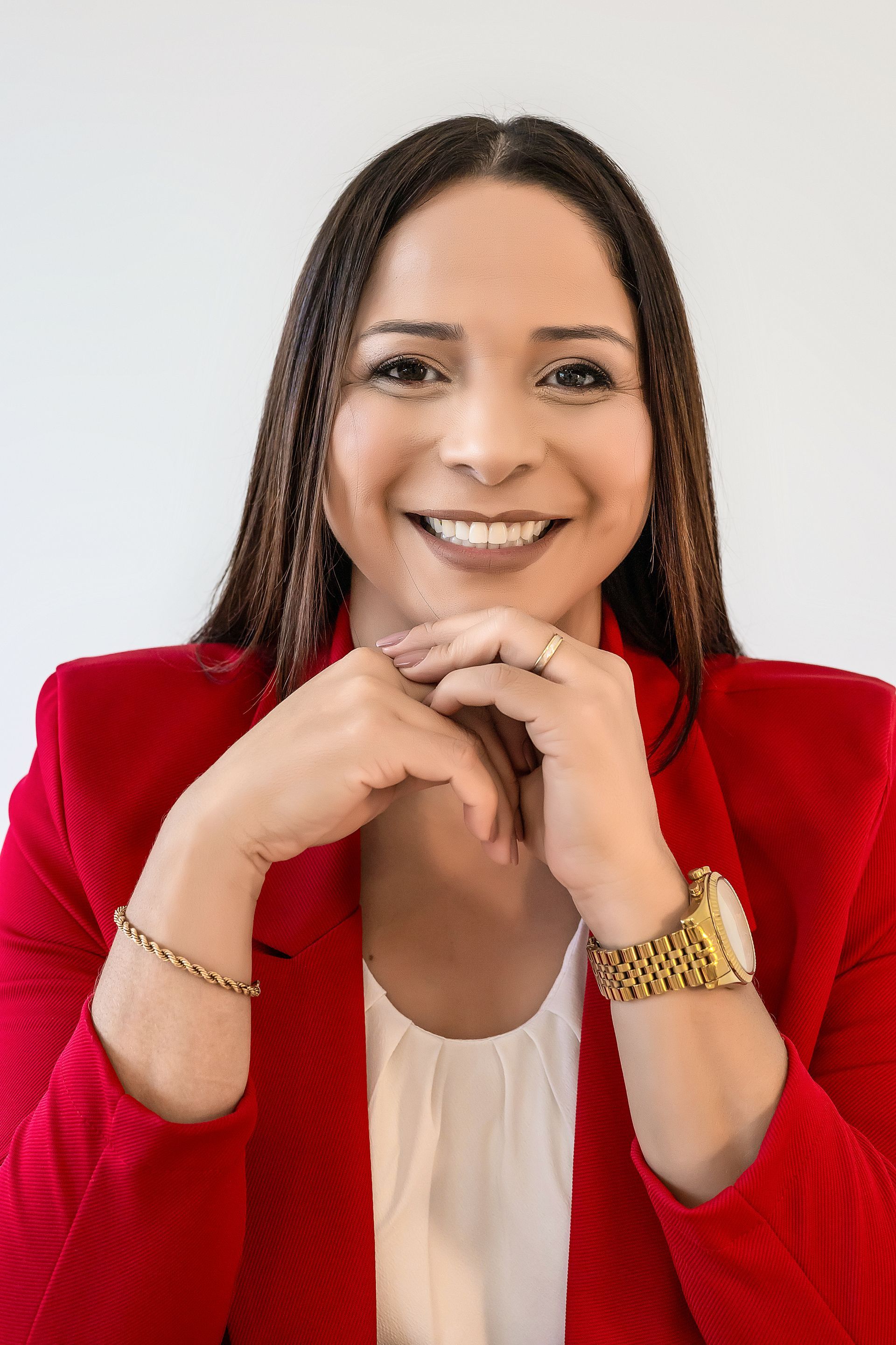 Mulher com cabelo trançado, vestindo um blazer vermelho, sorrindo com as mãos sob o queixo, relógio de ouro e joias.