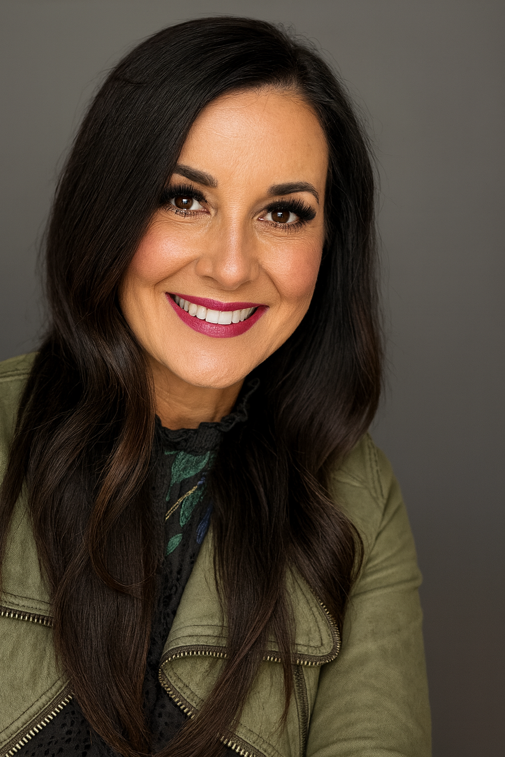 Woman with dark wavy hair smiles, wearing an olive green jacket, against a grey backdrop.