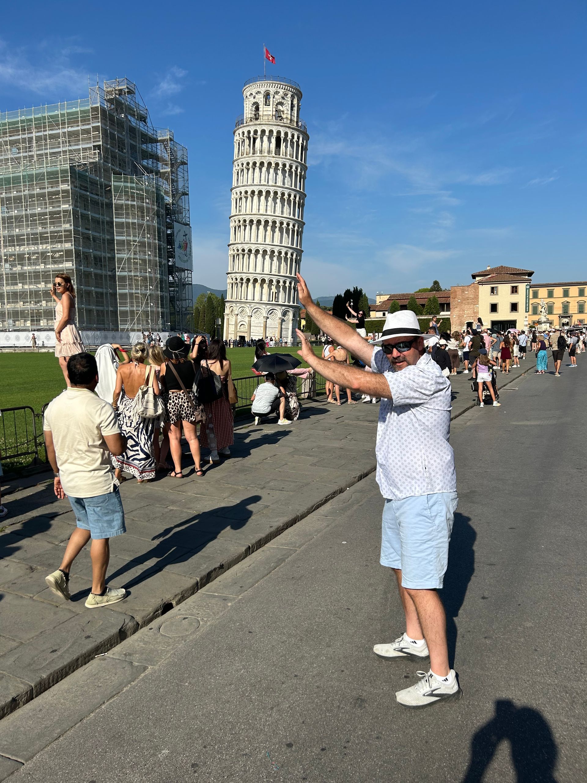 Man posing with his arms up, as if holding up the Leaning Tower of Pisa, with people in the background. Sunny day.