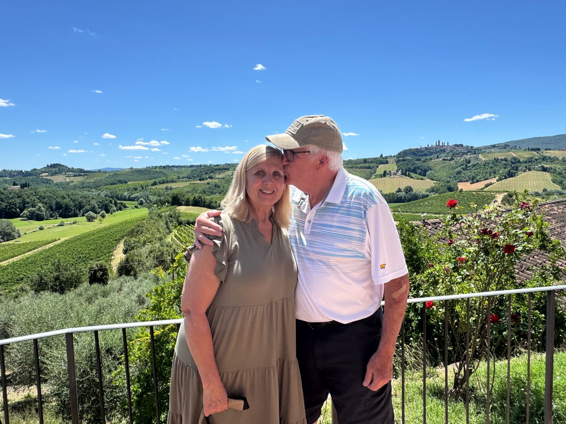 Man kisses woman's cheek; couple poses outdoors with scenic Tuscan hills in background.
