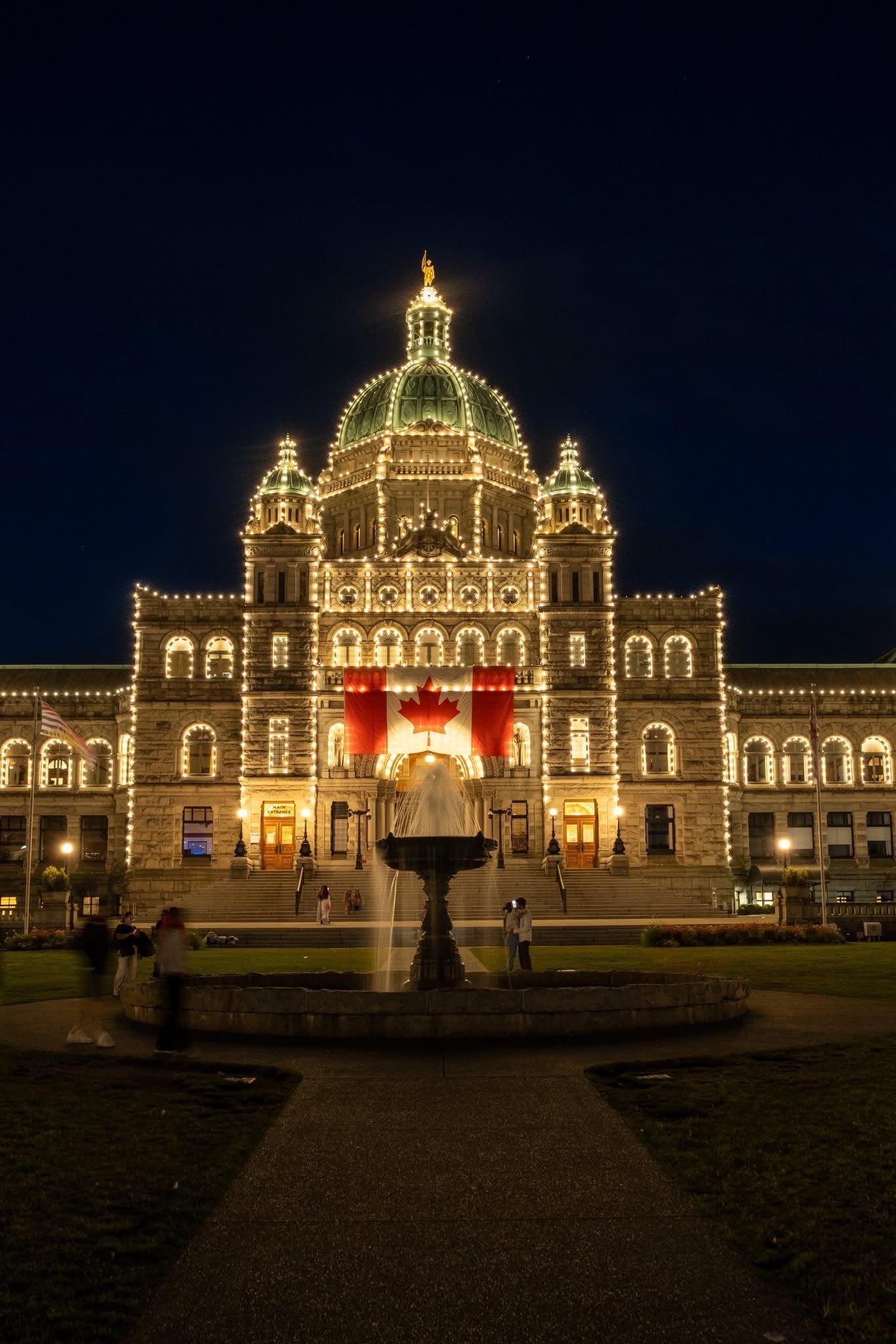 Parliament Buildings at night, illuminated with lights, Canadian flag, fountain in front.