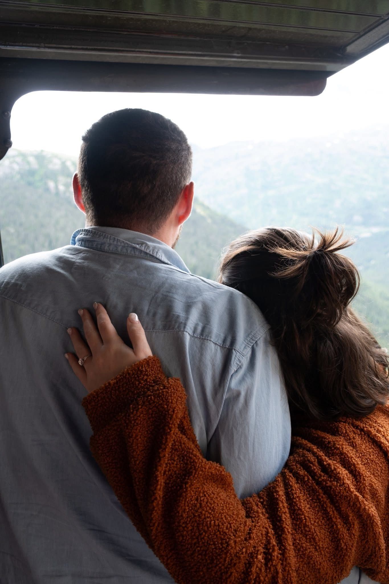 Couple embracing, looking at a mountain vista. Woman's arm around man's shoulders, wearing a brown sweater.