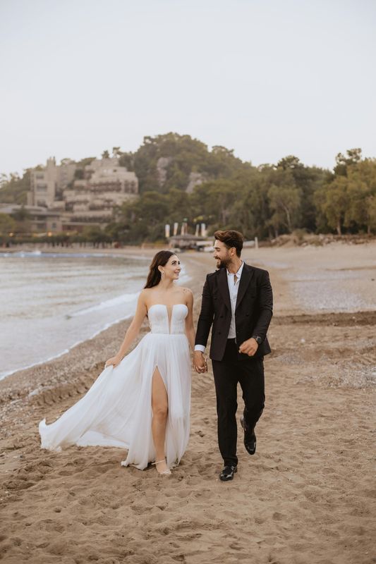 Couple in formal attire walking on beach, holding hands. Woman in white gown, man in black suit.