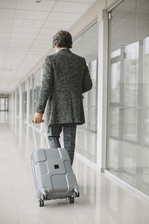 Person in patterned suit walking with rolling suitcase in a bright hallway.