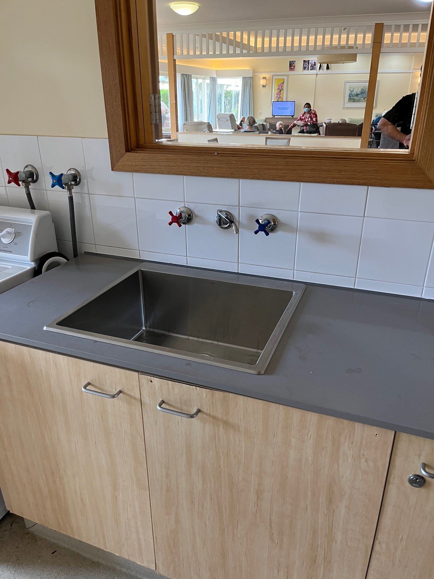 A stainless steel sink with faucets set into a gray countertop and wood-grain cabinet, with a mirror reflecting a room. — Distinct Plumbing Services In Kiama, NSW