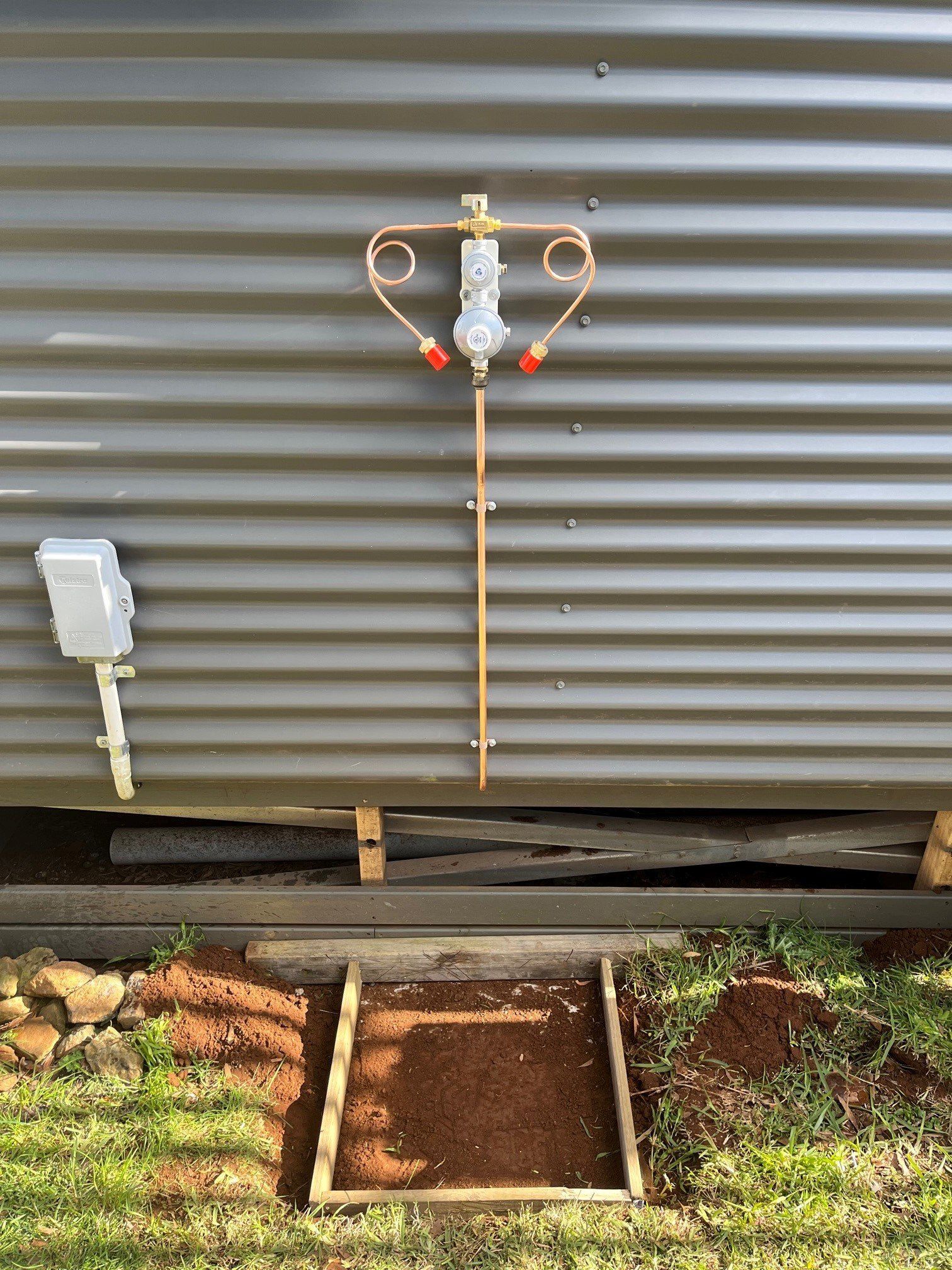A copper gas regulator assembly mounted on a corrugated gray wall above a framed, dirt-filled rectangular base.