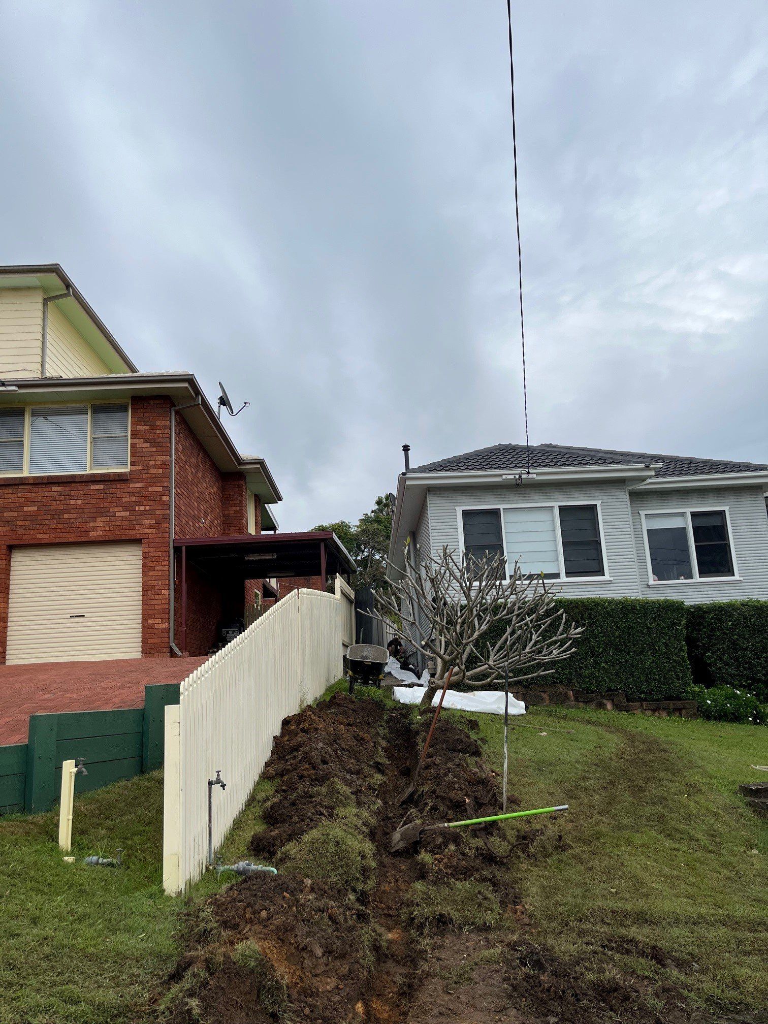 A freshly dug trench in a residential front yard between a brick house and a grey weatherboard house under a cloudy sky.