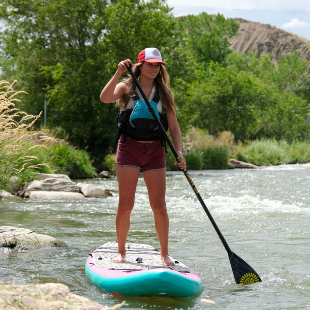 Woman paddleboarding on a river, holding a paddle, with green trees and rocky shore in the background