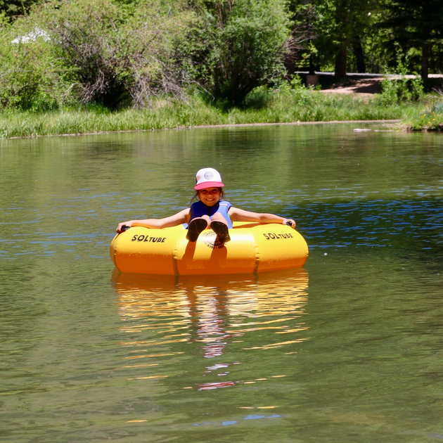Child in a pink helmet sitting on a yellow float in a calm river, with green trees in the background