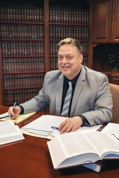Man in a suit seated at a desk, smiling, with books and papers in a library setting.