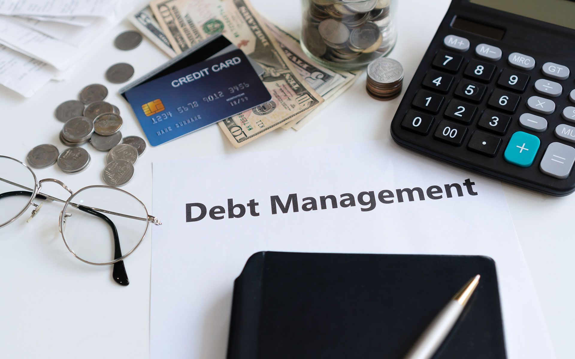 Debt management concept: credit card, cash, coins, calculator, glasses, and notepad on white surface.