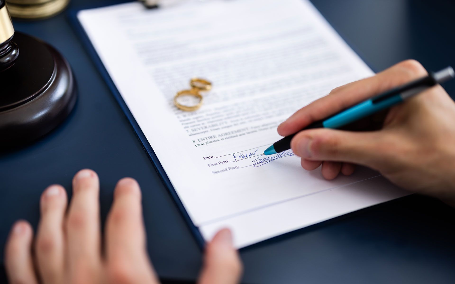 Person signing divorce papers at a desk, wedding rings nearby.