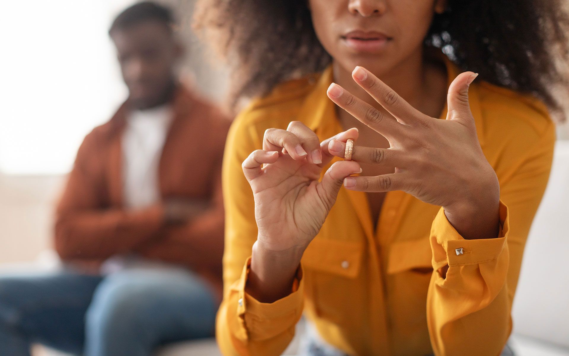 Woman removing wedding ring, man in background, signifying divorce.