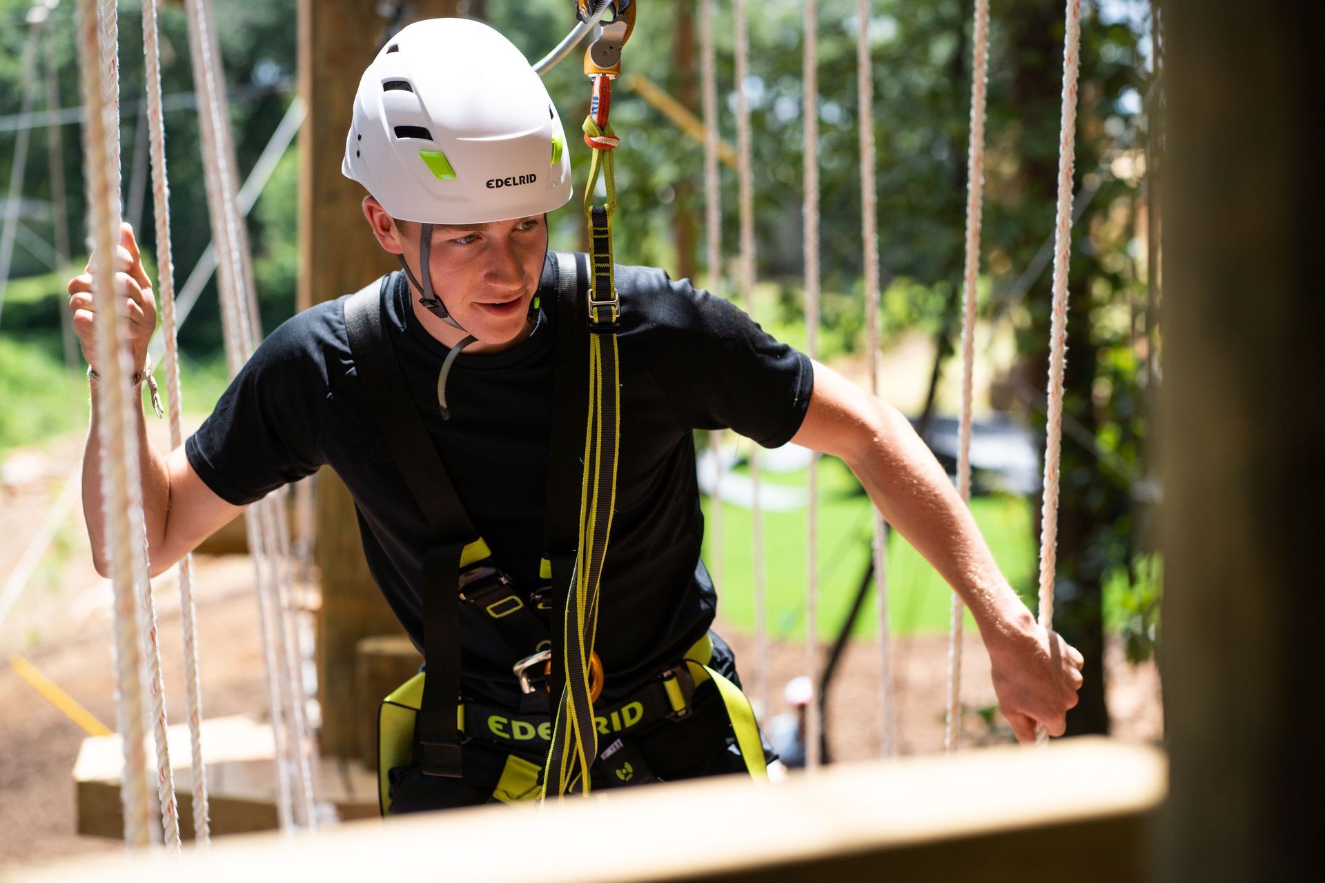 Teen in a harness and helmet navigating a treetop obstacle course, holding ropes, focused expression.