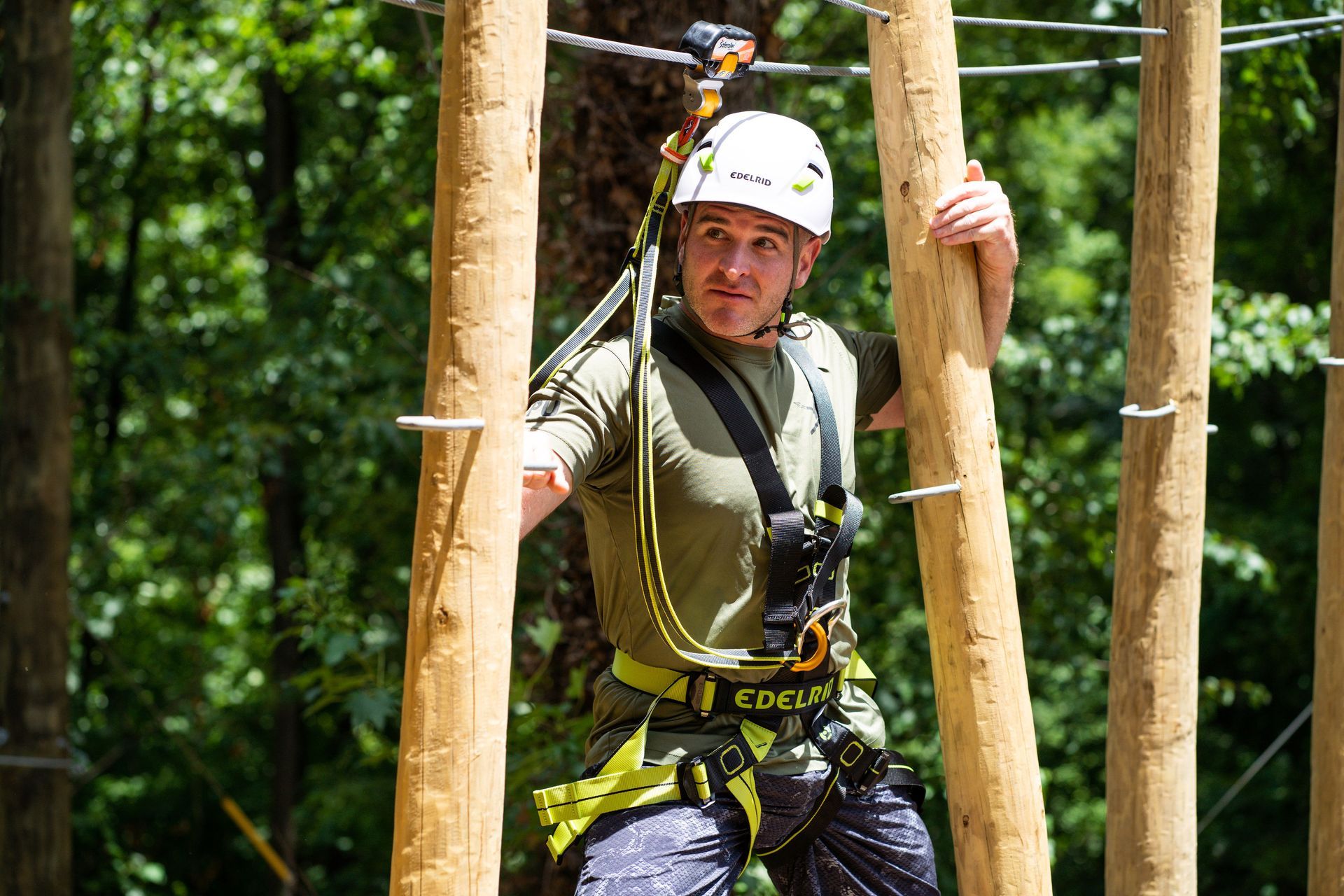 Man in safety gear on a treetop obstacle course, navigating between wooden beams. He wears a helmet and harness in a green forest setting.
