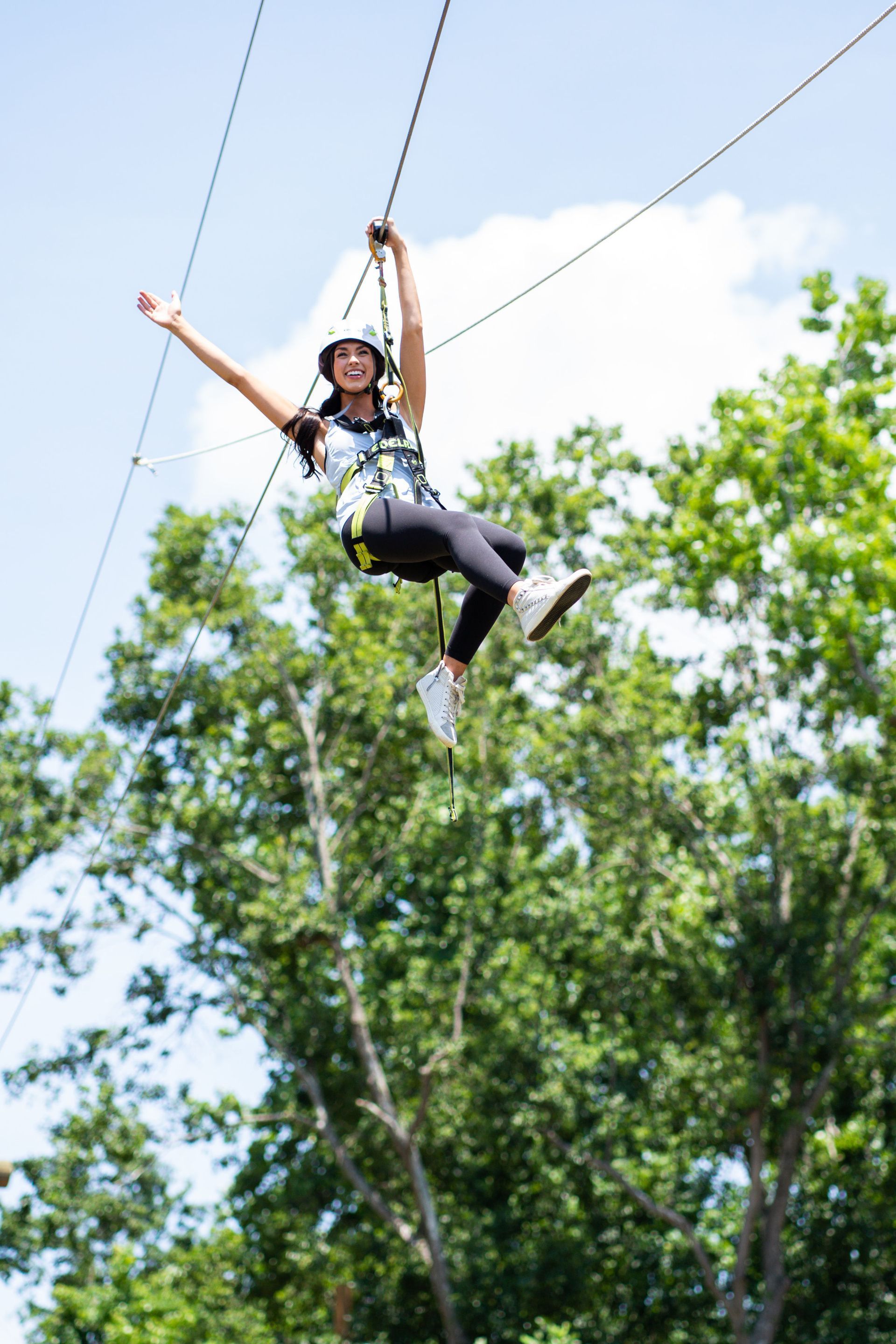 Woman ziplining through trees on a sunny day, arms outstretched. She wears a helmet and harness, smiling.
