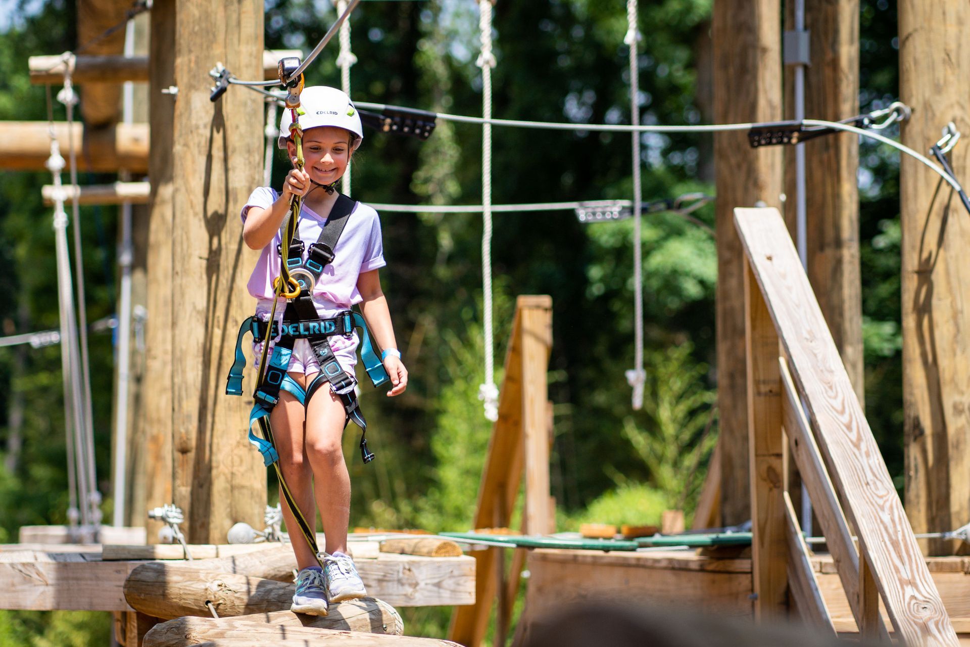 Young person in a harness and helmet smiles while walking across a ropes course, surrounded by wooden structures in a forest.