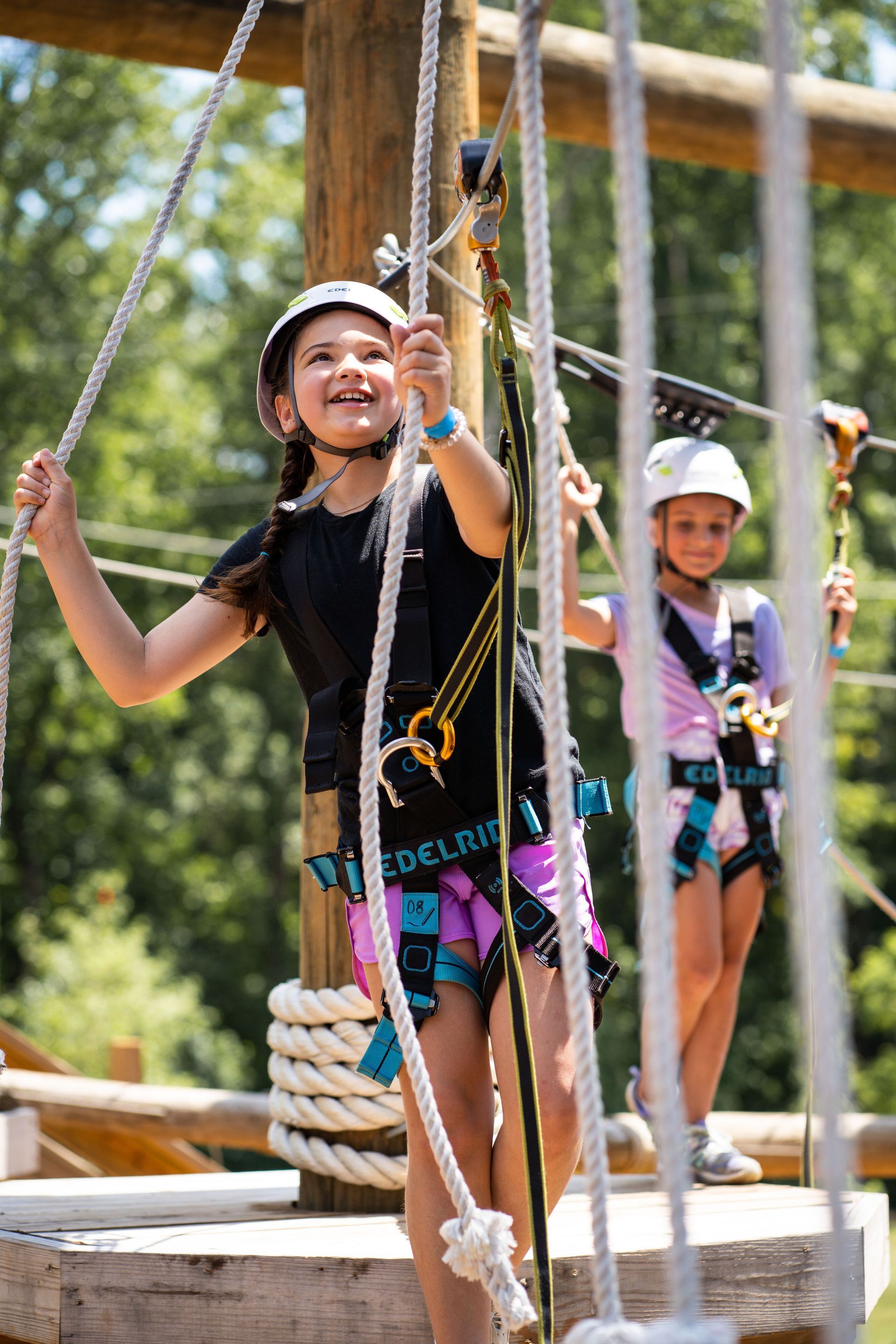 Two young girls wearing helmets and harnesses on a ropes course, smiling and navigating the course outdoors.