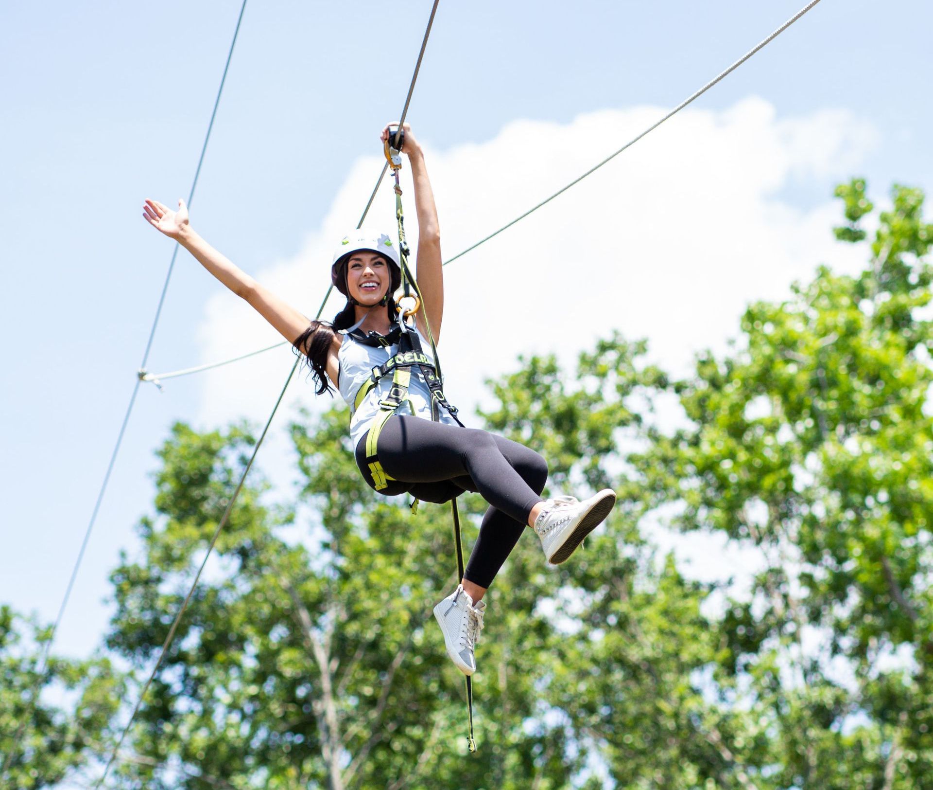 Woman ziplining with arms outstretched, smiling, wearing a helmet and harness, against a bright blue sky with trees.