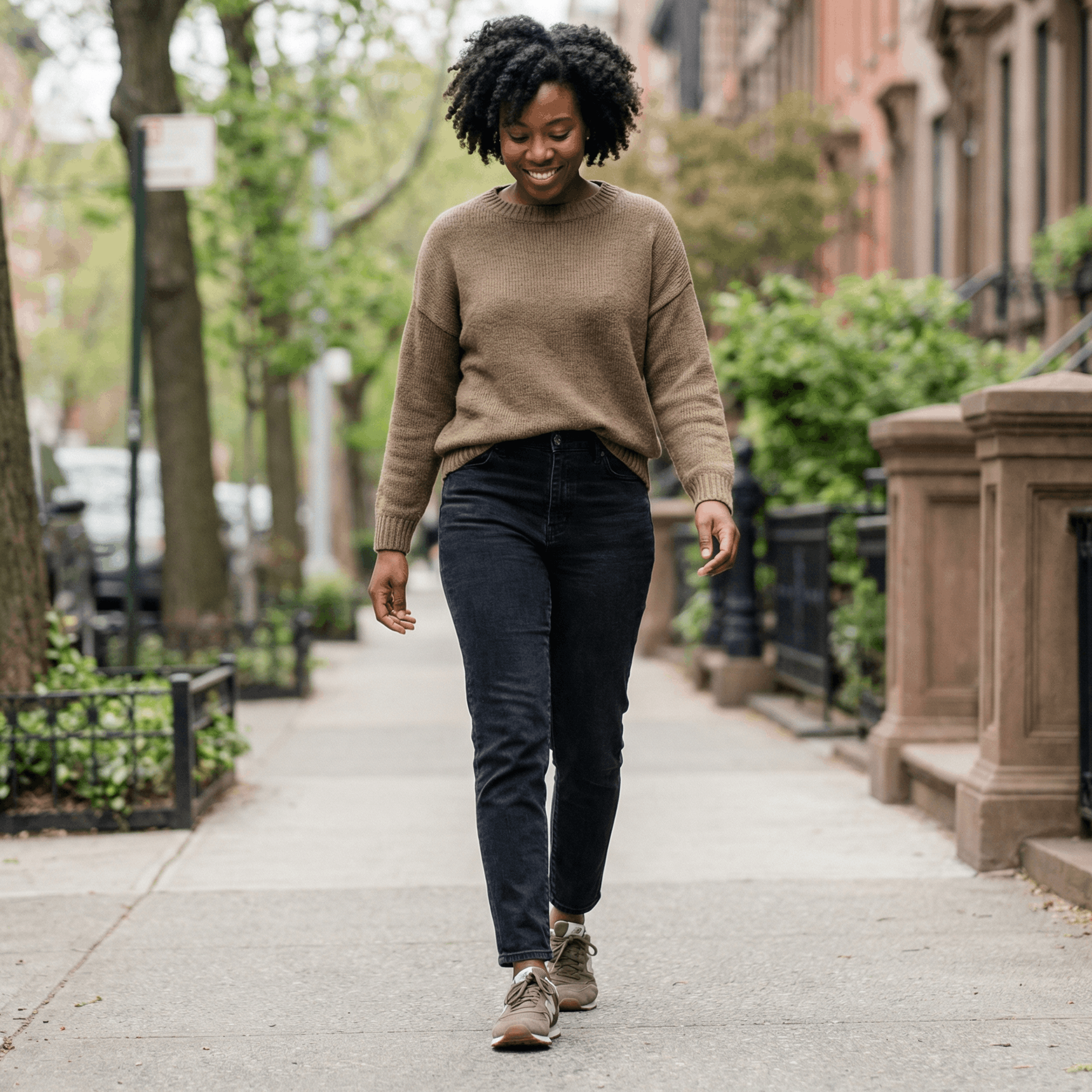 Woman in brown sweater and dark jeans walks smiling on sidewalk lined with trees and brownstone buildings.
