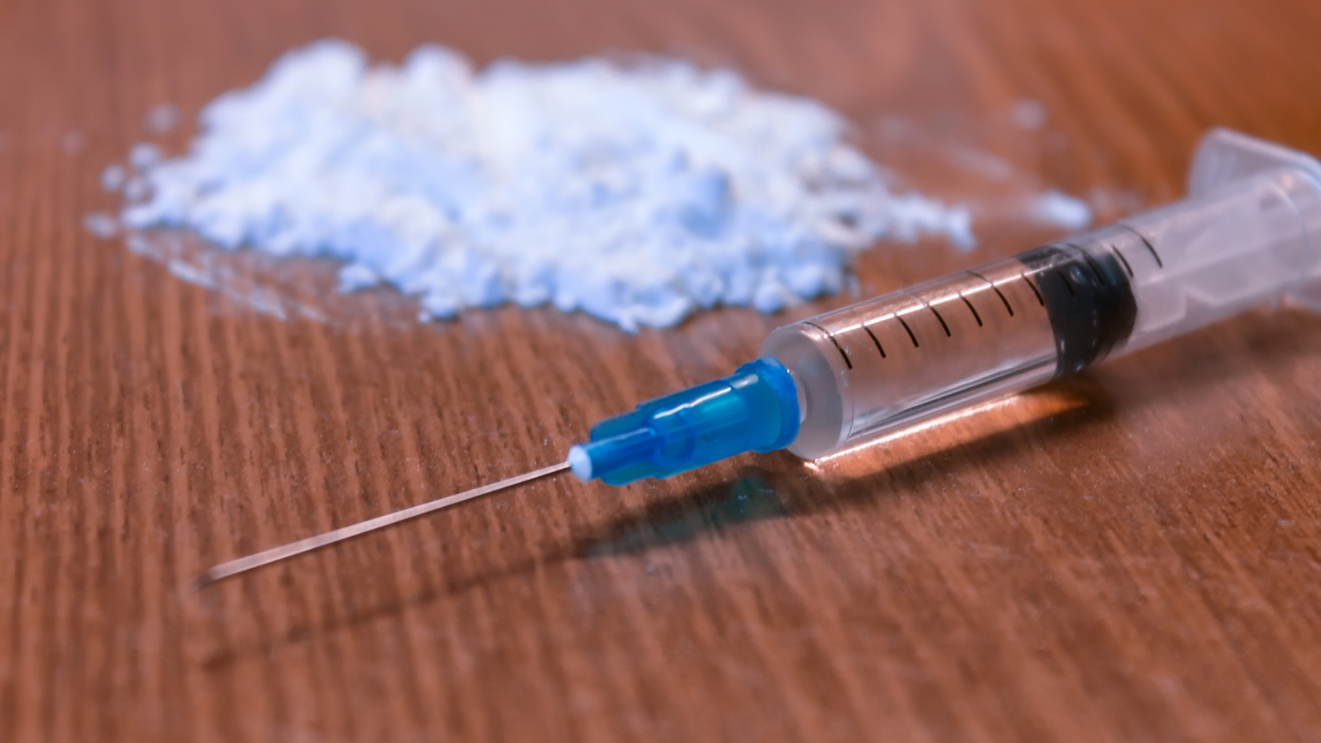 A syringe with a blue cap lies beside a pile of white powder on a wooden surface.