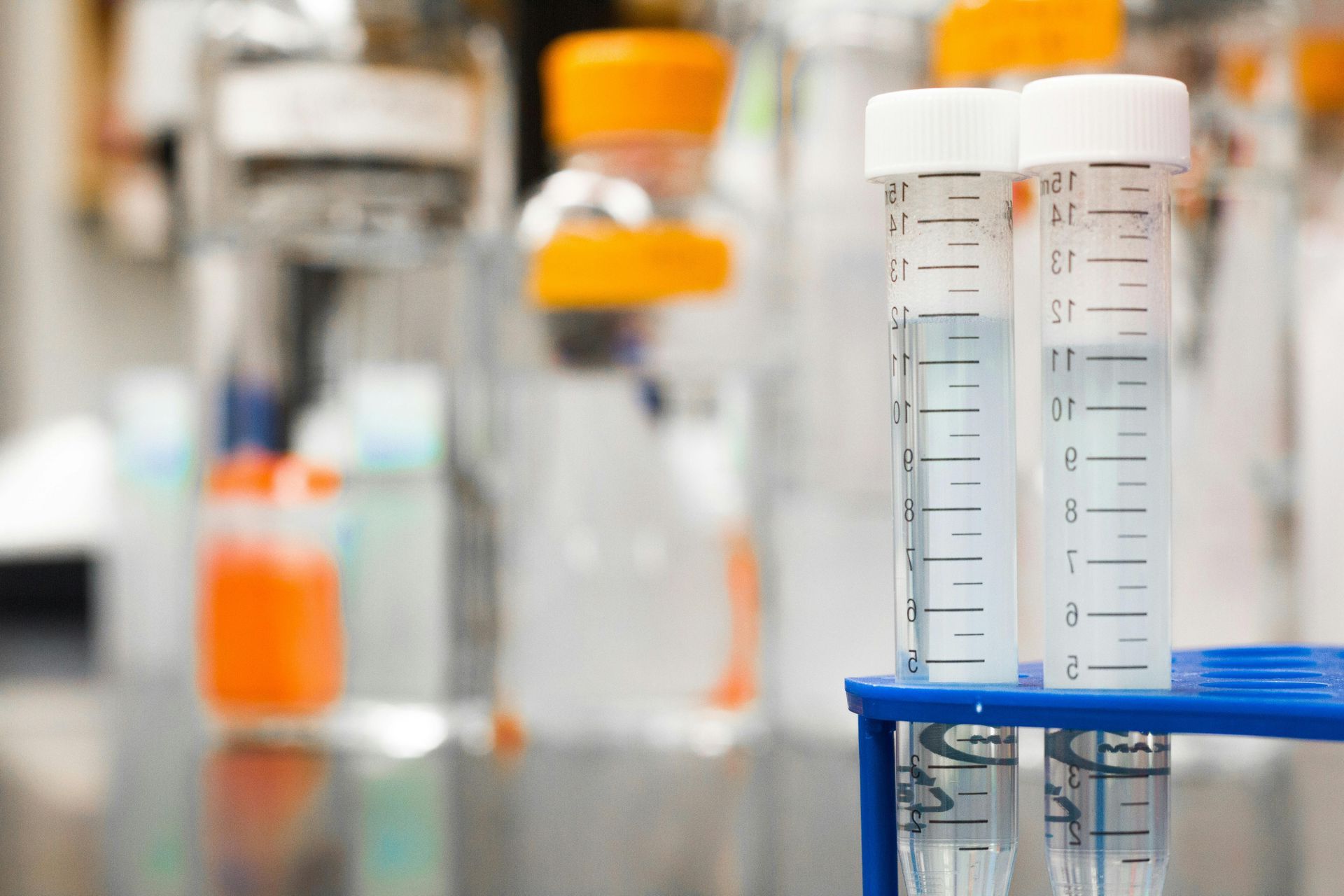 Test tubes with clear liquid in a lab rack, with blurred orange-capped vials in the background