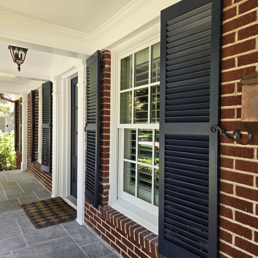 Black shutters on a brick building with white trim; front porch with a doormat.