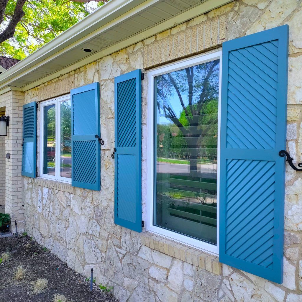 Blue shutters on a stone house. Windows reflect a tree and greenery.