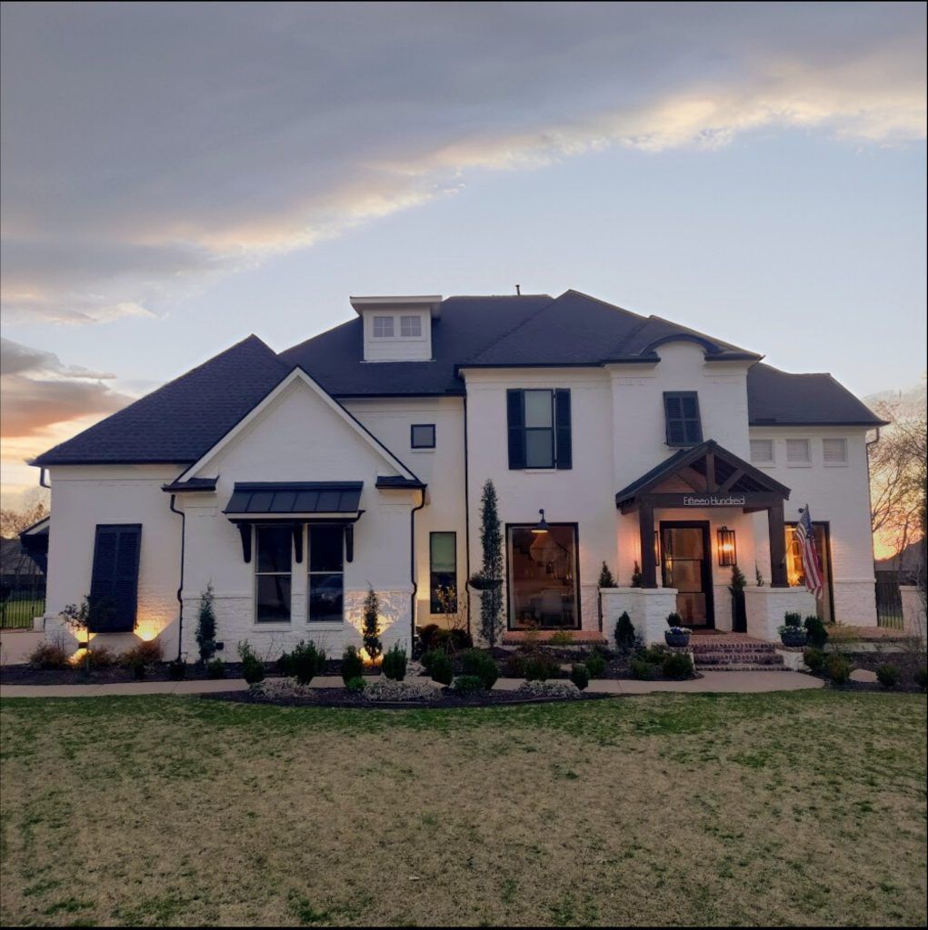 White brick house at dusk, with dark roof and trim. Lit landscaping and front porch.