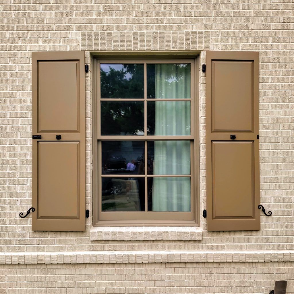 Tan shutters flanking a window with a beige frame set in a brick wall.