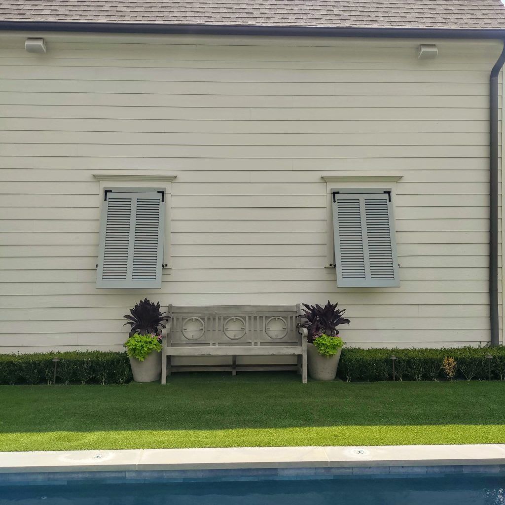 White house wall with two shuttered windows, a bench, potted flowers, and trimmed hedge above green lawn.