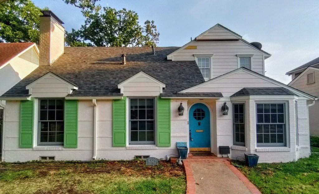 White house with green shutters, blue door, and red brick walkway.