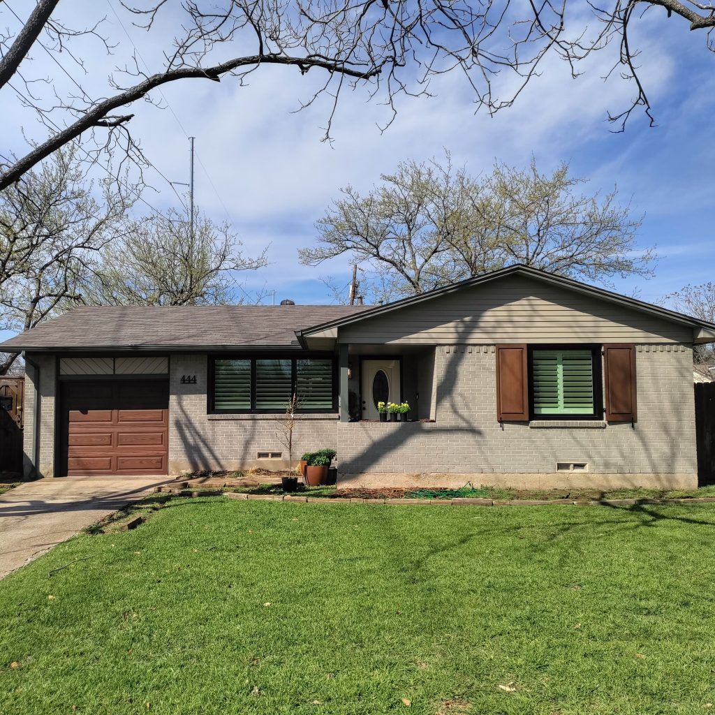 Ranch-style house with gray brick exterior, brown garage door and shutters, green lawn, and blue sky.