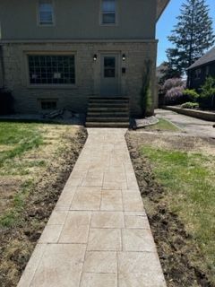 Concrete walkway leading to a house entrance with steps, bordered by grass and bare soil.