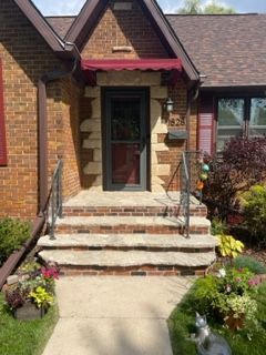 Brick house front entrance with steps, black railings, maroon awning, and flower beds along the walkway