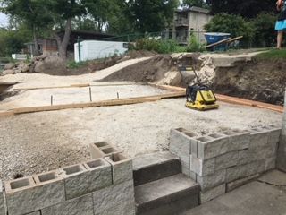 A yellow and black machine is sitting on top of gravel, cement blocks holding gravel in place to build a foundation. 