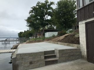 Concrete waterfront patio with steps beside a house, overlooking a calm lake and trees